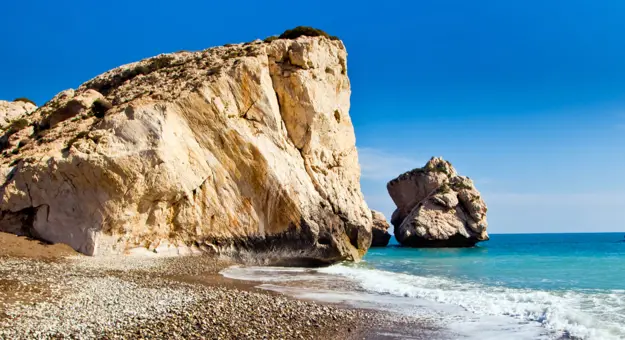 Large rock on a beach on the left with some smaller ones in front of it, the blue sea on the right, and pebbles and sand in the left forefront. 