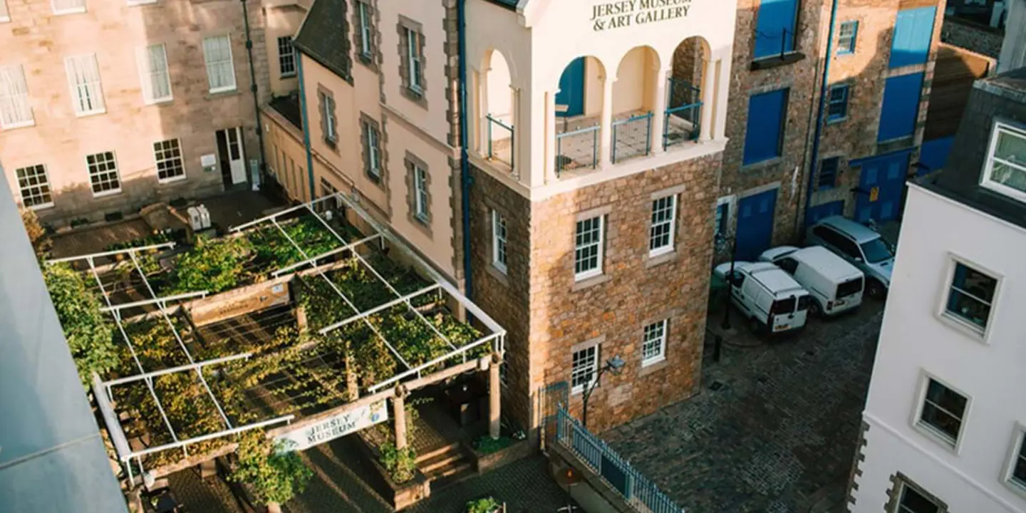 Bird's eye view of the Jersey Museum and Art Gallery, showing an open roof with plants tied to it and the Jersey Museum sign at the front of it to the left, and a car park with some parked white vehicles to the right