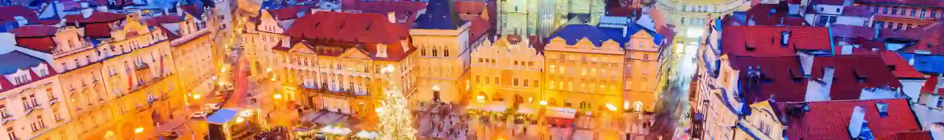 High angle shot of Christmas market in the town square of Prague, showing a lit up gothic church with two towers and spiky turrets, a monument with a circular barrier around it in the centre of the square, next to a large lit up Christmas tree. White roofs of the stalls are all around the square surrounded by people.