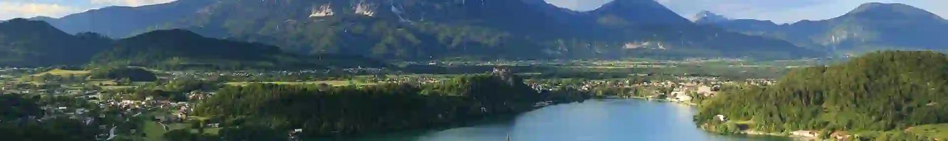 Wide angle shot of Lake Bled, showing its forested surrounding areas, and mountains in the distance. In the middle of the lake, there is a small piece of land with trees and a towered building.